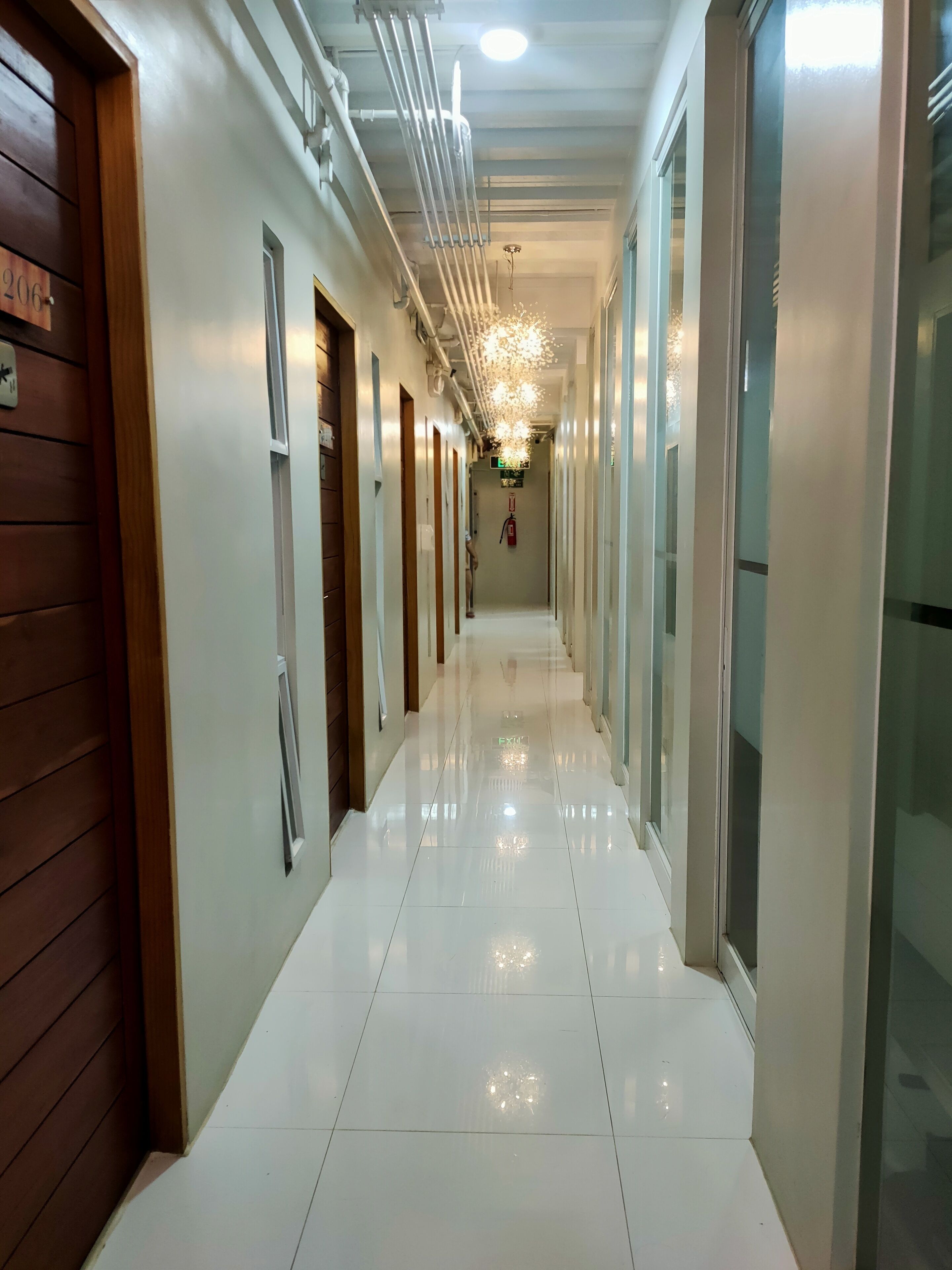 Un pasillo de hotel con pisos de baldosas blancas, puertas de madera y un techo con tuberías a la vista y una lámpara colgante.