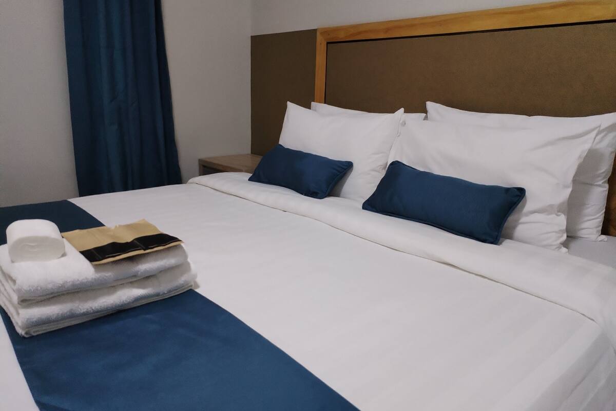 A neatly made bed with white and blue bedding, a wooden headboard, and a small bedside table with a lamp.