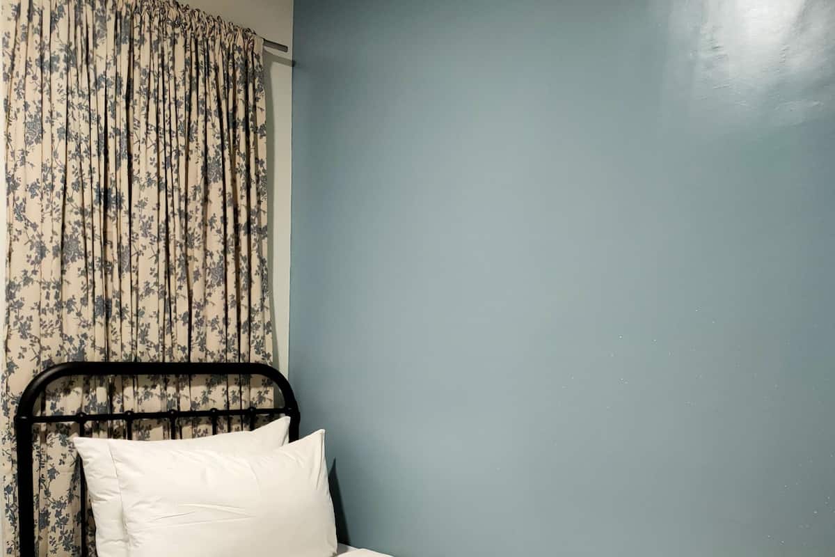 A single bed with a white mattress and pillows, a black metal frame, a floral curtain, and a blue wall.