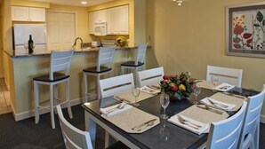 Un coin repas avec une table dressée pour quatre personnes, des chaises blanches, un vase de fruits et, au fond, une cuisine.