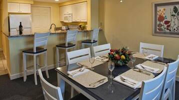 A dining area with a table set for four, white chairs, a vase of fruit, and a kitchen in the background.