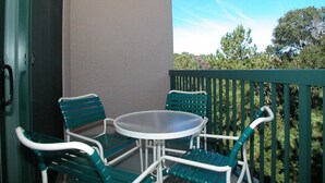 Un balcon avec une table ronde et deux chaises, une porte et une vue sur les arbres et le ciel.