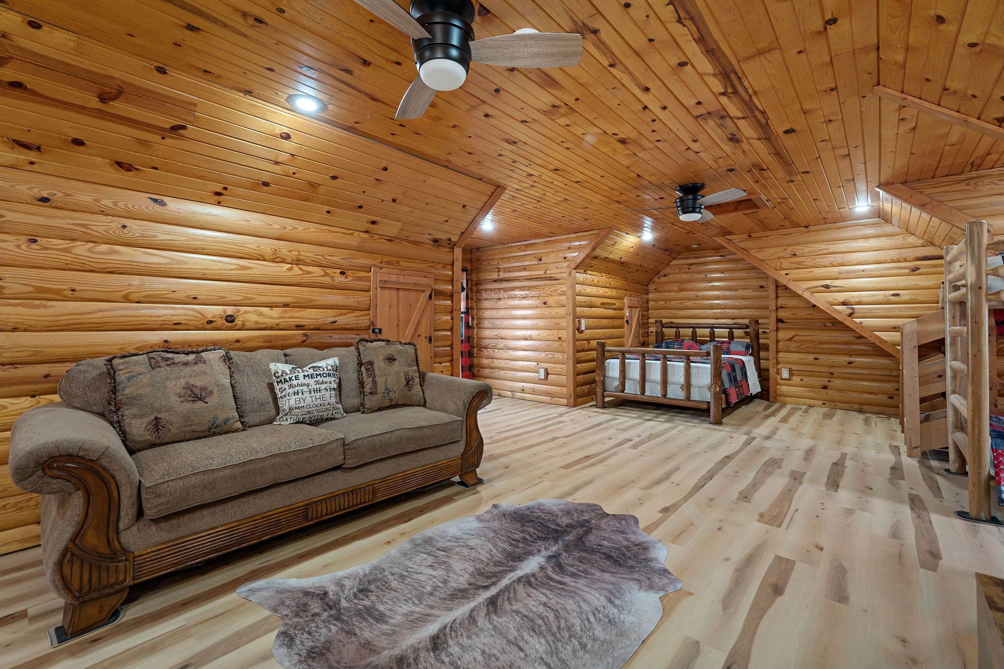 Interior de una cabaña de madera con piso de madera, un sofá, un manto de piel y una cama con una manta roja.