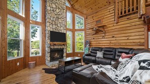 Interior de una cabaña de troncos con chimenea de piedra, sofá de cuero y ventanas amplias con vistas al exterior.