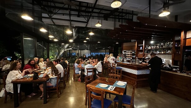 A restaurant with wooden tables and chairs, a bar area, and a large window with greenery visible outside.