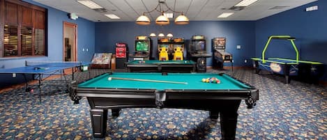 Mesa de bilhar com bolas, mesa de snooker e jogos de fliperama em um ambiente com carpete estampado.