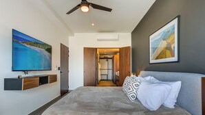 Ein modernes Schlafzimmer mit einem großen Bett, einem Deckenventilator, einem an der Wand montierten Fernseher und einem Flur zum Badezimmer.