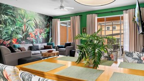 Ein modernes Wohnzimmer mit einer großen tropischen Wandmalerei, ein Essbereich mit einem Holztisch und grünen Matten sowie ein Blick auf ein Gebäude durch große Fenster.