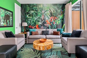 A living room with a green wall, a mural of tropical foliage, a grey sofa set, and a round wooden coffee table.