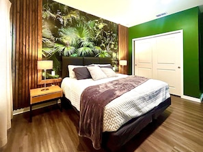 A bedroom with a bed, a nightstand, a lamp, a wall with a plant mural, and a door.