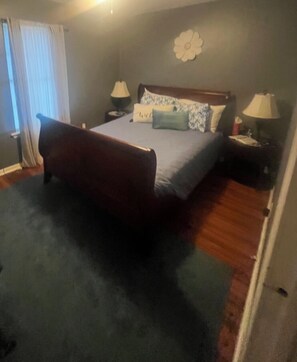 A bedroom with a bed, two bedside tables, a nightstand, a window with curtains, and a wall decoration.