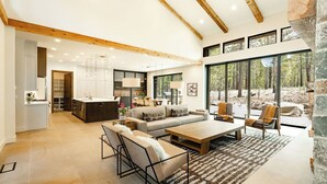 A spacious living area with a large sofa, coffee table, and chairs, a kitchen with modern appliances, and large windows offering a view of the outdoors. - Sable Rock Retreat- Modern Luxury w/ 5 beds, 6 ba, spa, sauna, 2nd living room. (Bend)