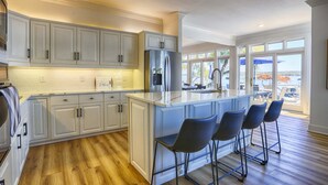 A modern kitchen with white cabinetry, a stainless steel refrigerator, and a large island with bar stools. - Lakeview Heaven - Elegant Canandaigua Lake Getaway | Dock + Hot Tub (Canandaigua)