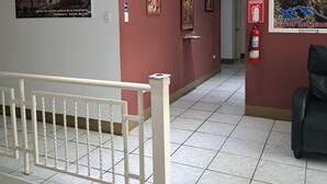 Un pasillo con barandilla blanca, paredes rojas y piso de baldosas. Se observan cuadros enmarcados en las paredes, un extintor y una silla negra.