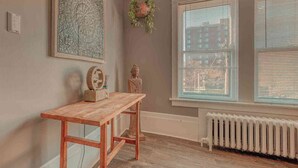 A room with a wooden table, a hanging plant, a Buddha statue, a wall tapestry, and a window with blinds. - Historic Riverfront Tropical Gem w/ River Views (Harrisburg)