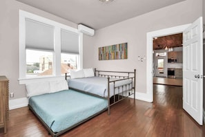 Een slaapkamer met een bed, een raam met zonwering, een schilderij en uitzicht op een keuken.