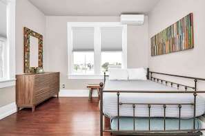 Une chambre avec un lit en métal, une commode en bois, une grande fenêtre avec des stores et un tableau abstrait aux couleurs vives accroché au mur.