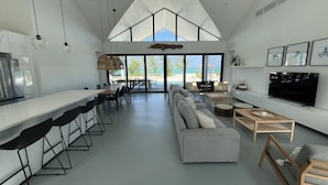 Une cuisine moderne dotée d’un long comptoir et de tabourets de bar, un coin repas avec un grand canapé et un téléviseur, et une vue sur la plage grâce à de grandes fenêtres.