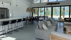Un vaste espace de vie comprenant un grand canapé gris, une table basse en bois et un coin repas avec une longue table et des chaises. Une cuisine moderne avec un réfrigérateur en acier inoxydable est visible au fond.