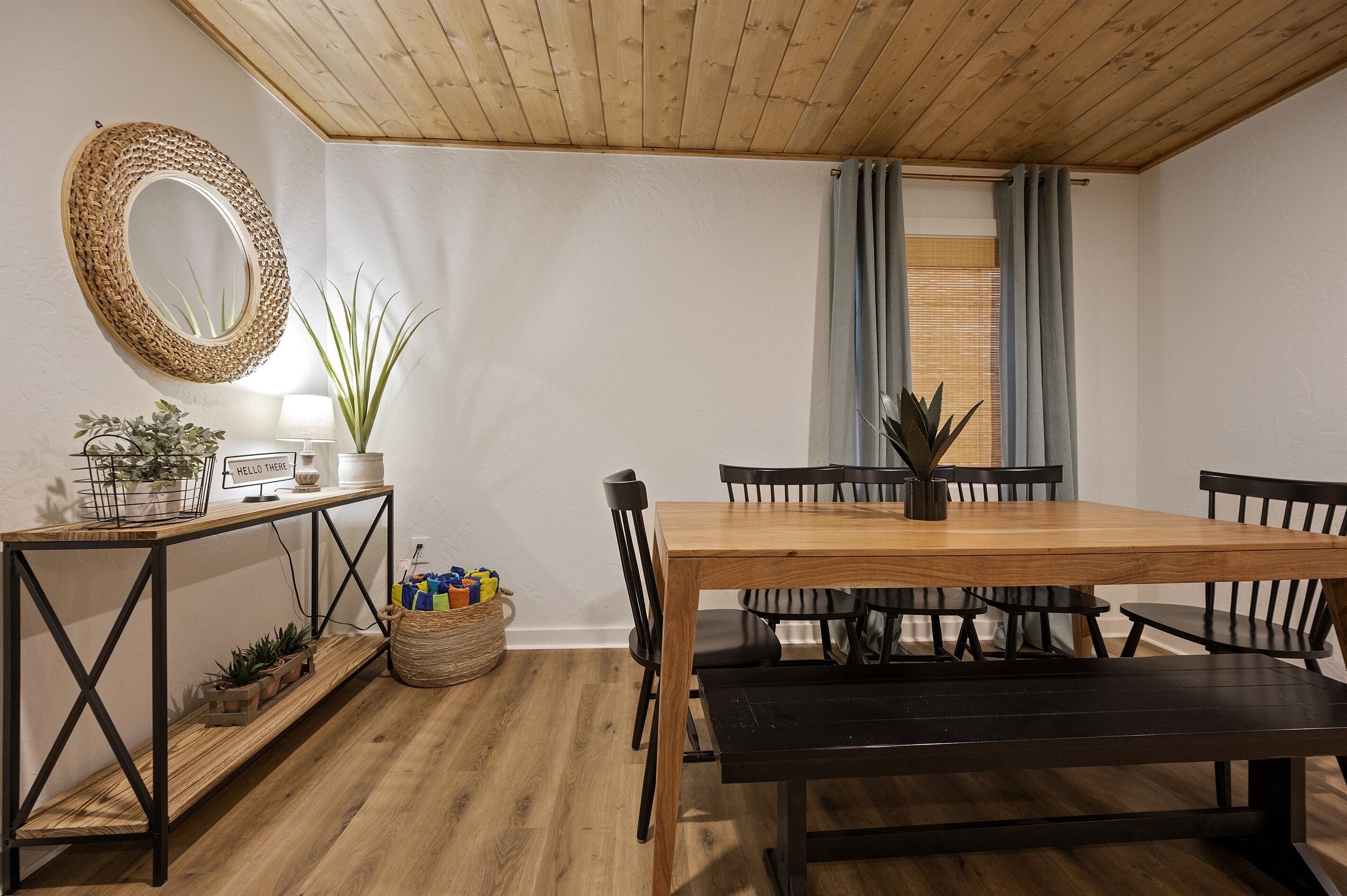 Et spiseområde med et træbord, sorte stole, en bænk, et vævet spejl og et konsolbord med planter og en lampe.
