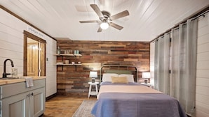Ein Schlafzimmer mit Holzkopfteil, einem Bett mit blauer Decke, Nachttischlampen, einem kleinen Tisch mit Lampe, Holzboden und Deckenventilator.