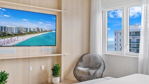 Ein modernes Hotelzimmer mit einem großen Fernseher, der einen Strand zeigt, einem bequemen Sessel, einem Bett und einem Fenster mit Blick auf das Meer und die Gebäude.