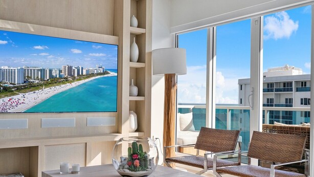 Ein modernes Hotelzimmer mit einem großen Fernseher, der einen Strand zeigt, einem Glastisch mit Sukkulenten als Dekoration und einem Balkon mit Korbstühlen.