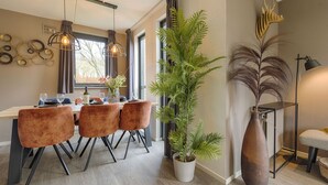 A dining area with a table set for four, surrounded by velvet chairs, a tall potted plant, and a decorative wall piece.