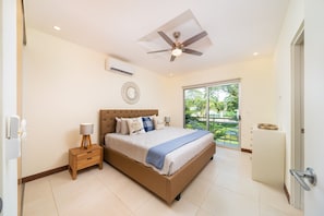 A bedroom with a bed, bedside table, lamp, ceiling fan, air conditioning unit, and a view of greenery through a sliding door.