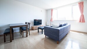 A modern living room with a wooden dining table, chairs, a sofa, a flat-screen TV, and a window with curtains. - LAVISH (Kamifurano)