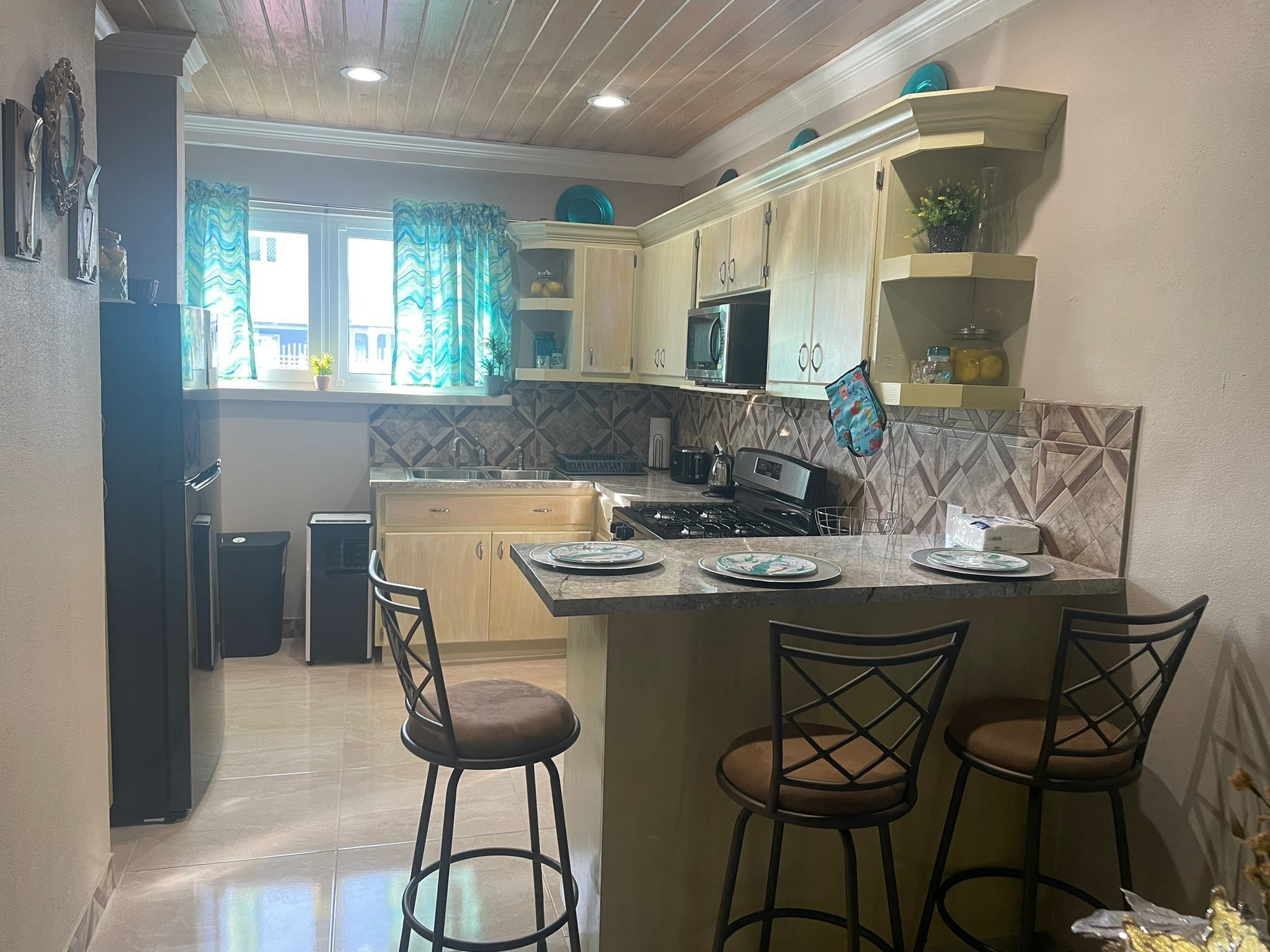 Una cocina con armarios de madera, un microondas, una estufa y un lavamanos. Hay dos taburetes de bar y una ventana con cortinas.