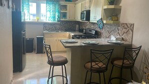Cozinha com armários de madeira, micro-ondas, fogão e pia. Há duas banquetas de bar e uma janela com cortinas.