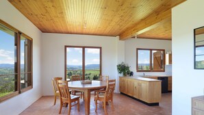 Une cuisine spacieuse dotée de meubles en bois et d’un grand îlot central, un coin repas avec une table ronde et des chaises, et de grandes fenêtres offrant une vue sur la campagne.