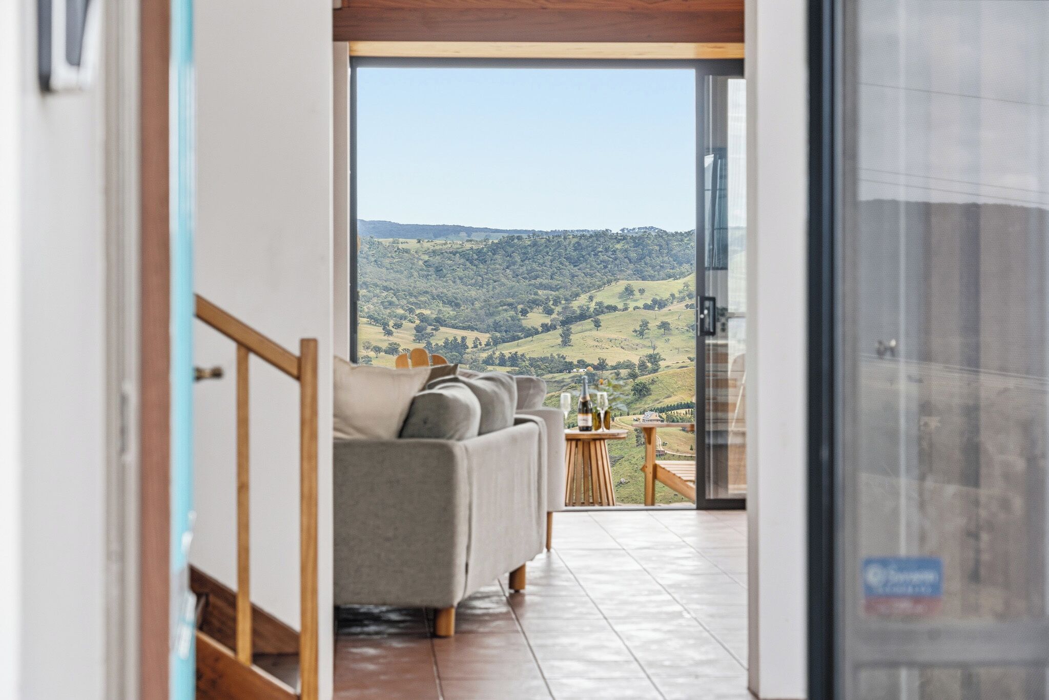 A modern interior with a sofa and a small table, a glass door leading to a balcony with a view of a hilly landscape.