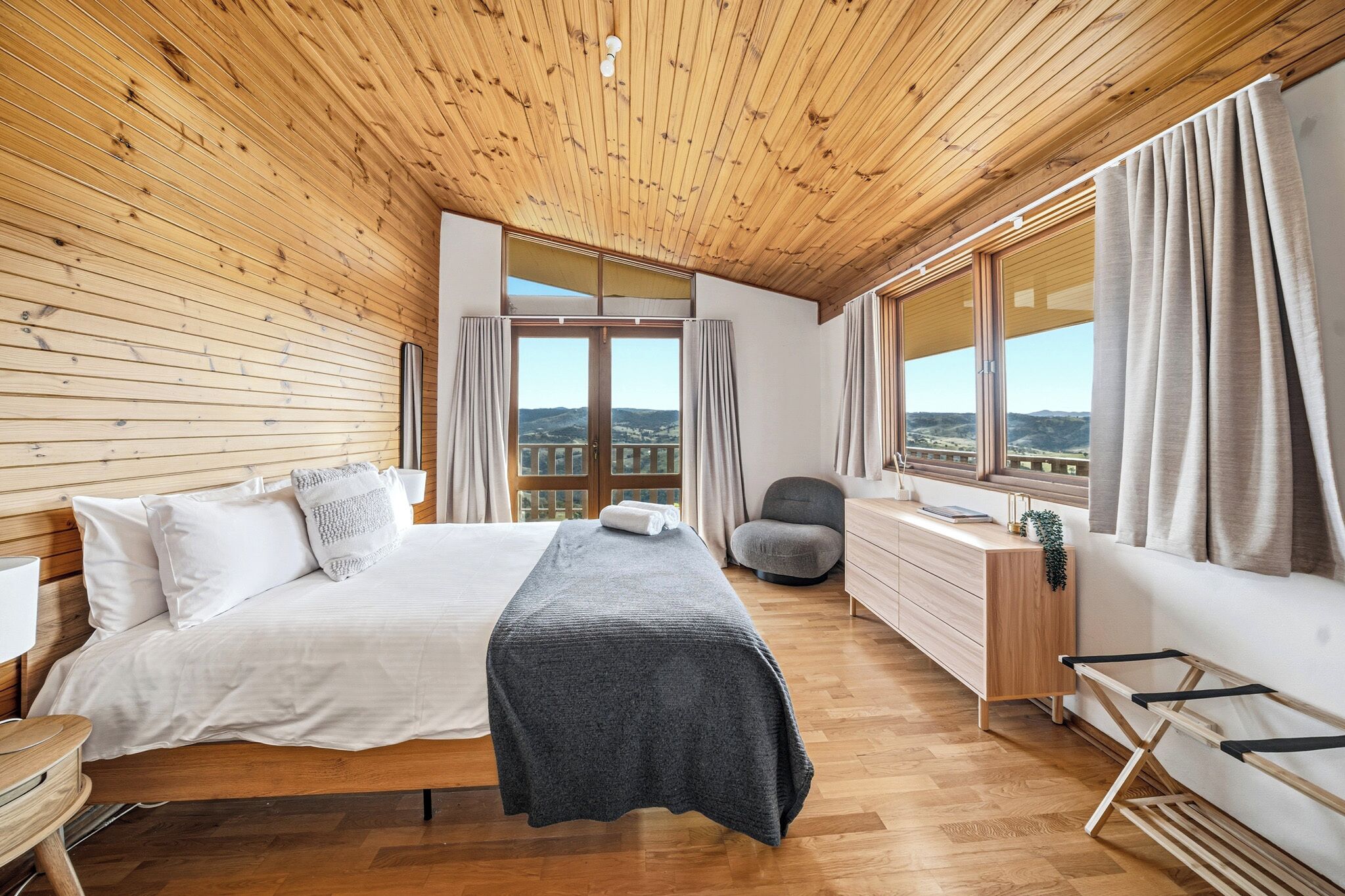 A bedroom with a wooden ceiling, a bed, a desk, and a view of the outdoors.