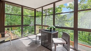 A sunlit room with large windows, wicker furniture, and a view of a forested area.