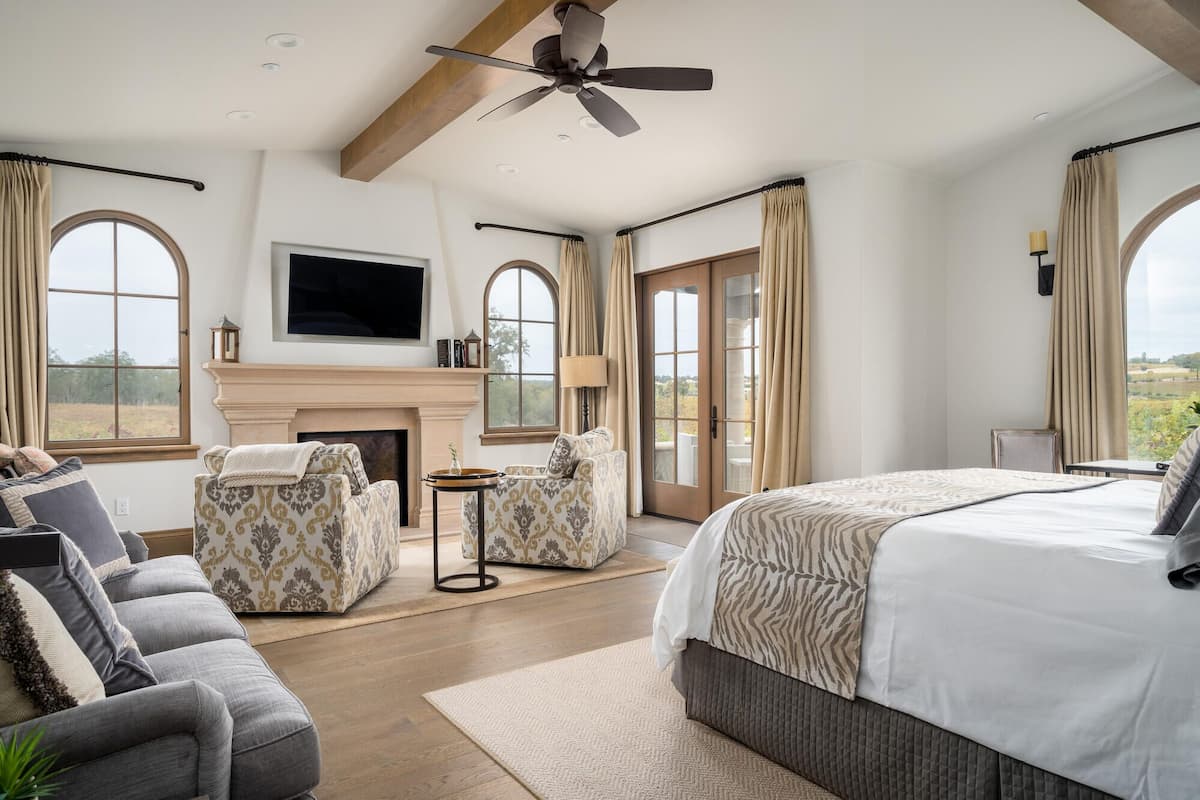 A spacious bedroom with a large bed, two armchairs, a fireplace, and a TV mounted on the wall.