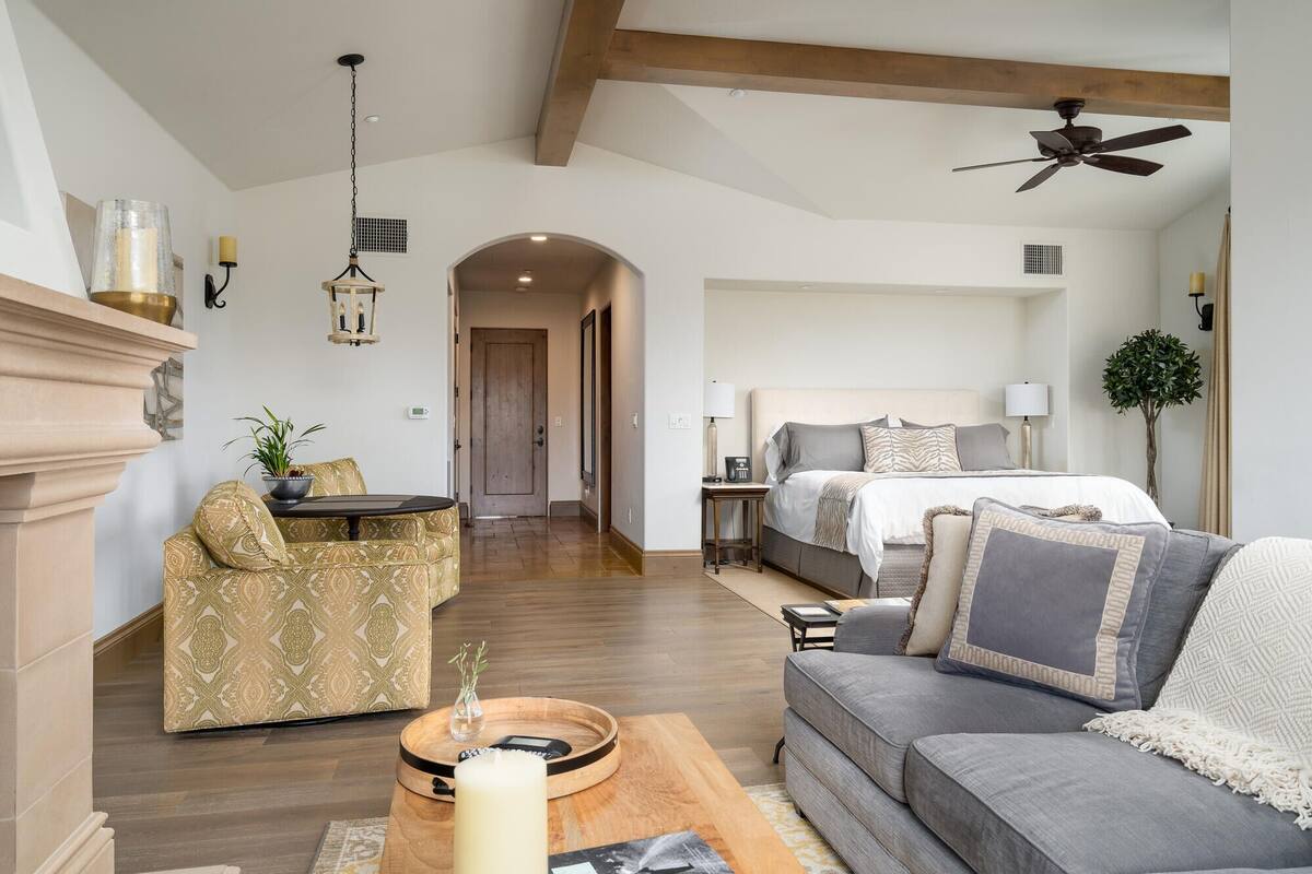 A spacious living room with a sofa, a coffee table, and a dining area. There is a bed in the background with a headboard and a bedside table.