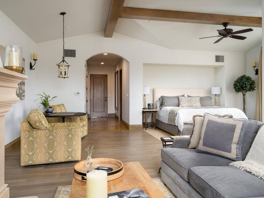 A spacious living room with a sofa, a coffee table, and a dining area. There is a bed in the background with a headboard and a bedside table.