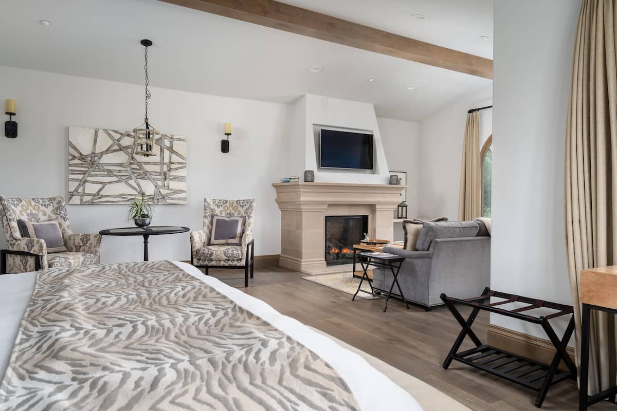 A modern hotel room with a fireplace, a large bed, a seating area with chairs, and a television.