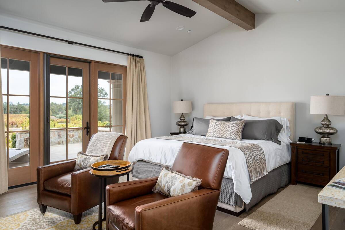 A bedroom with a large bed, two bedside tables, a chair, and a view of a vineyard through large windows.
