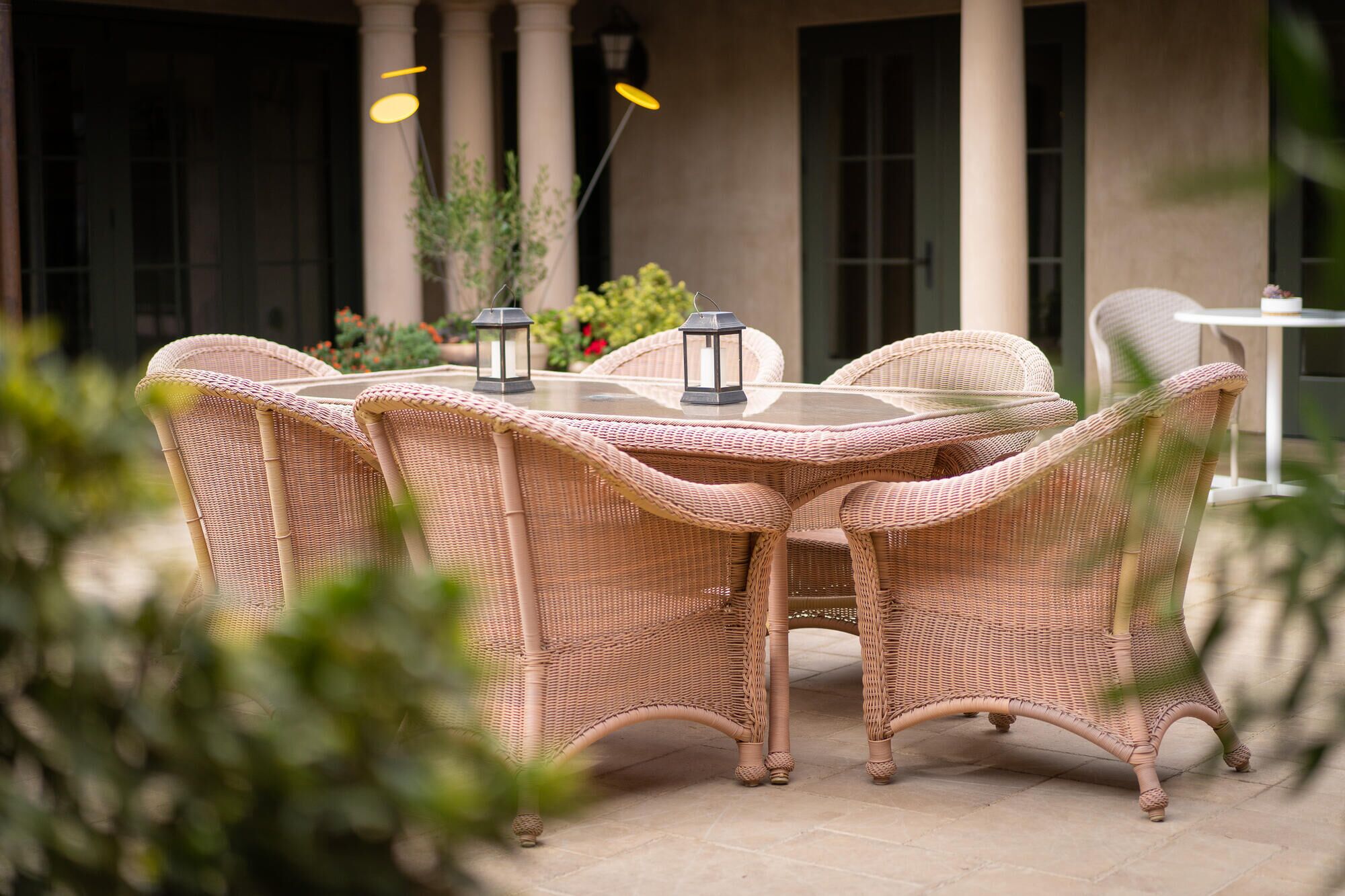 A wicker outdoor dining set with a glass table and two lanterns, surrounded by columns and greenery.