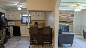A modern kitchen with white cabinets, a microwave, and a coffee maker. There are two bar stools and a gray sofa in the living area. - Riverfront Home (Tidioute)