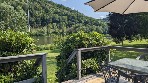 A deck with a table and chairs overlooking a lake, surrounded by lush greenery and a forested hillside. - Riverfront Home (Tidioute)