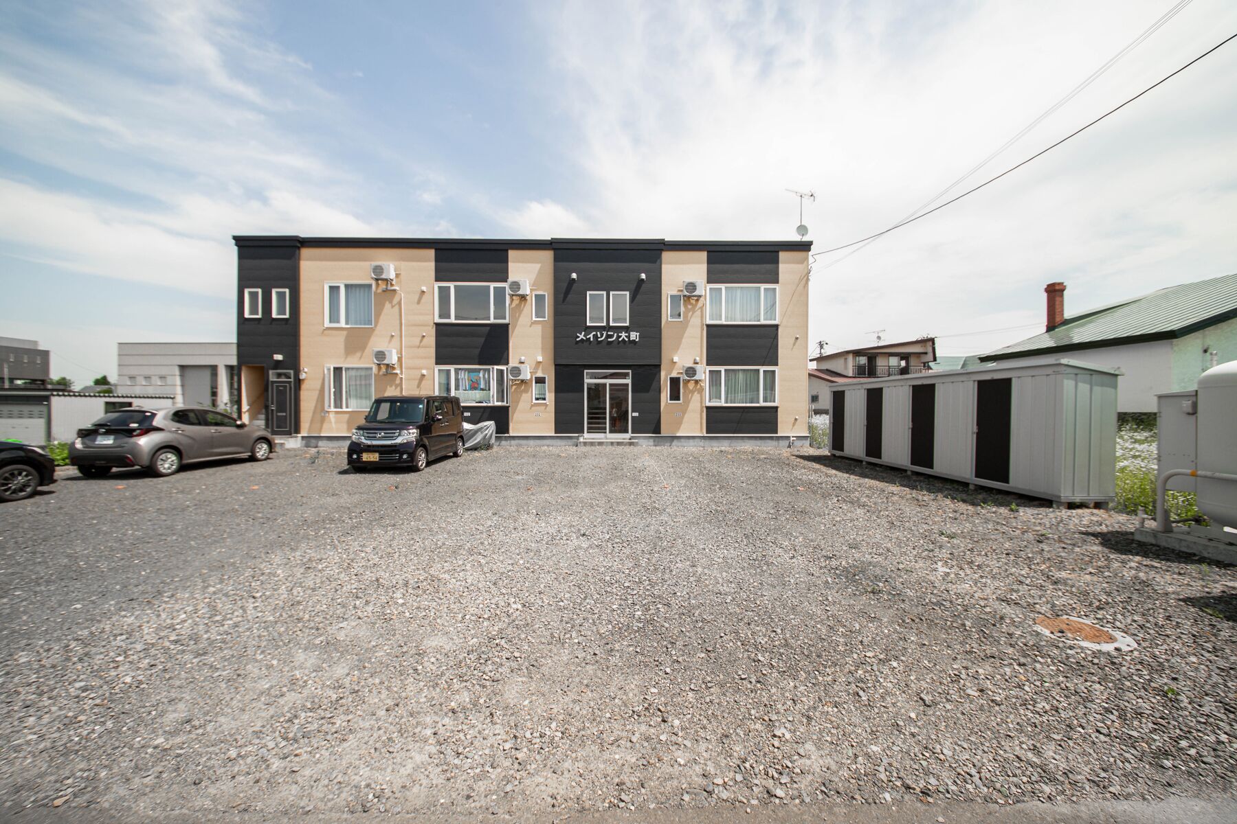 A multi-story building with a black and beige facade, a covered parking area, and a gravel driveway.