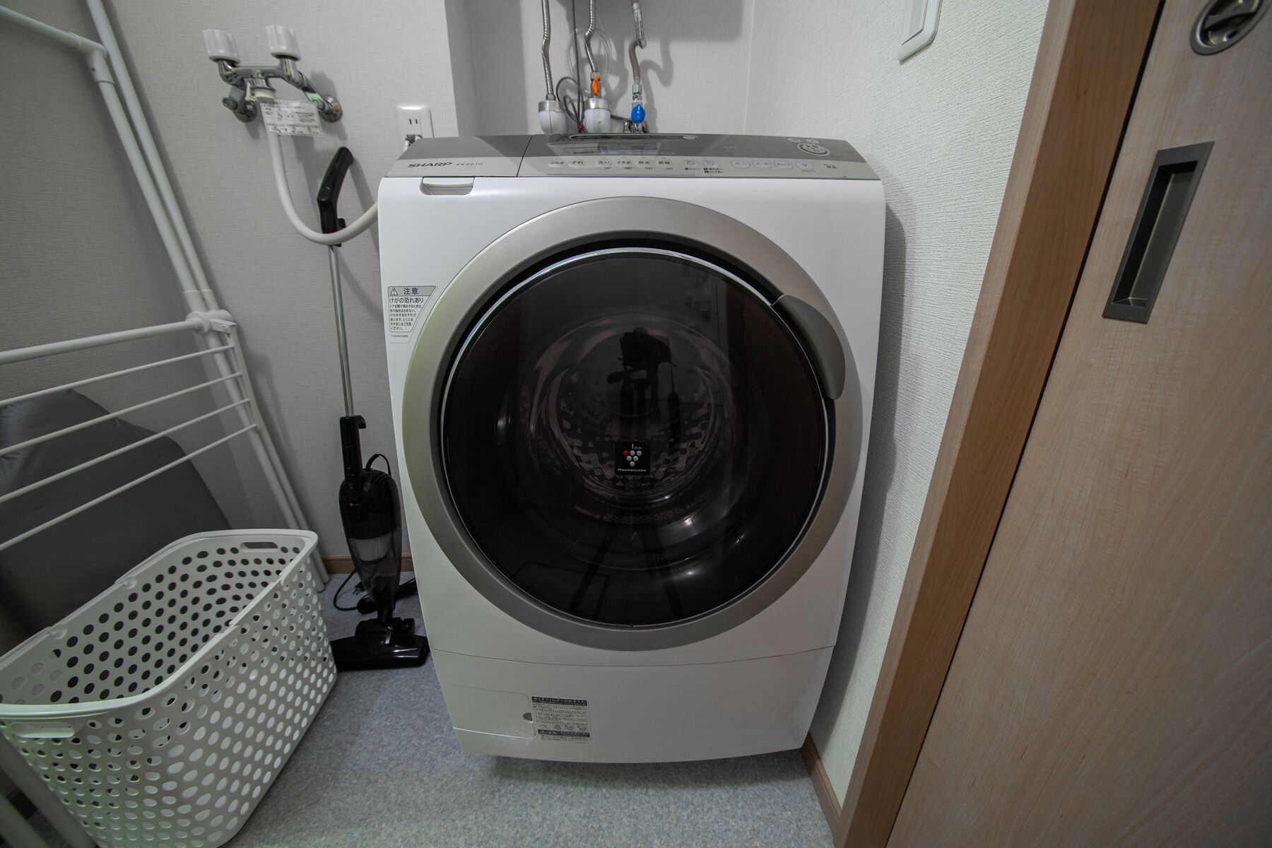 A compact, modern washing machine in a small laundry area with a metal rack and a plastic basket.
