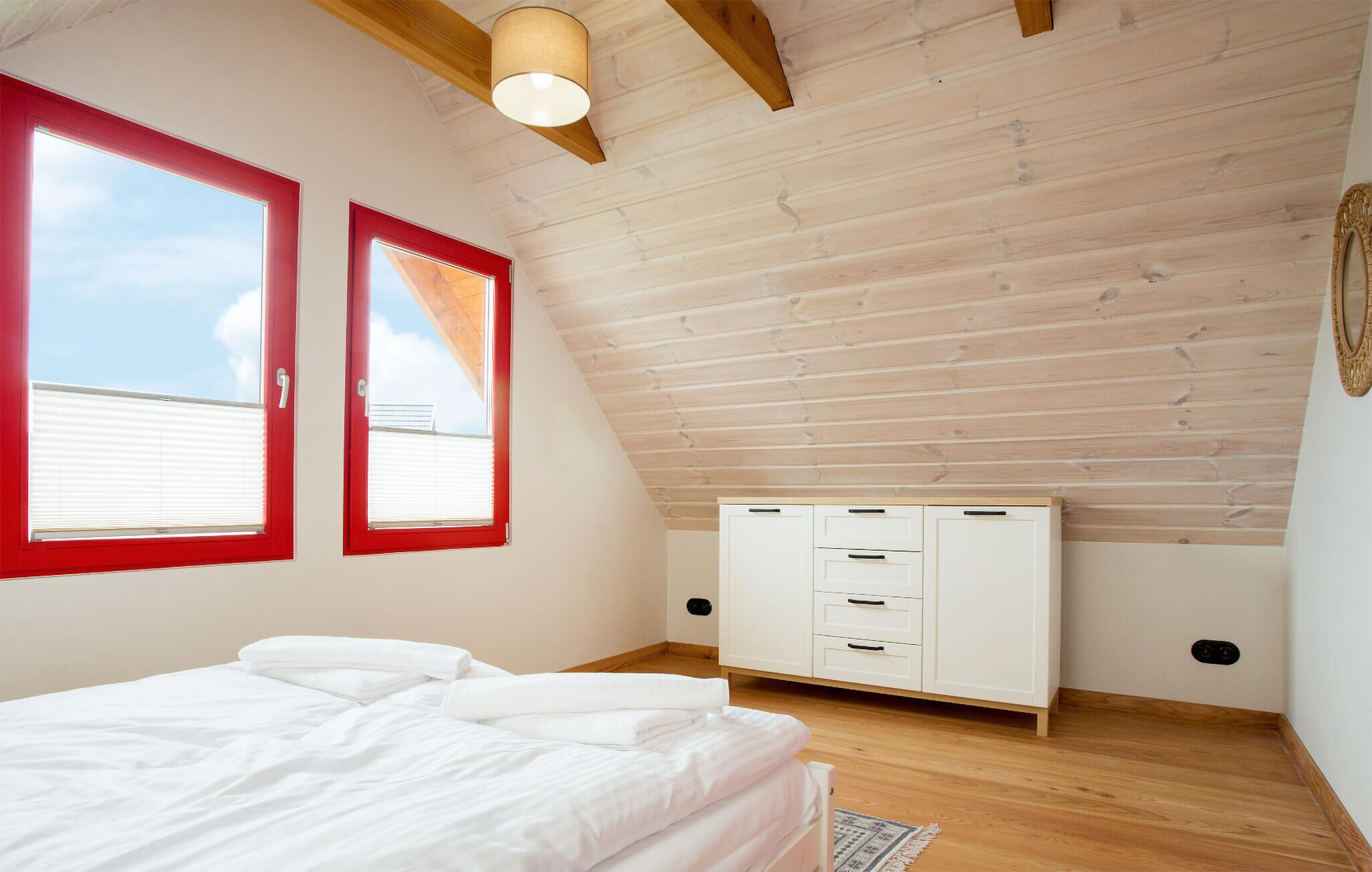 Une chambre avec un plafond en pente, des poutres en bois, un lit avec une literie blanche, deux fenêtres à cadre rouge et une armoire blanche.