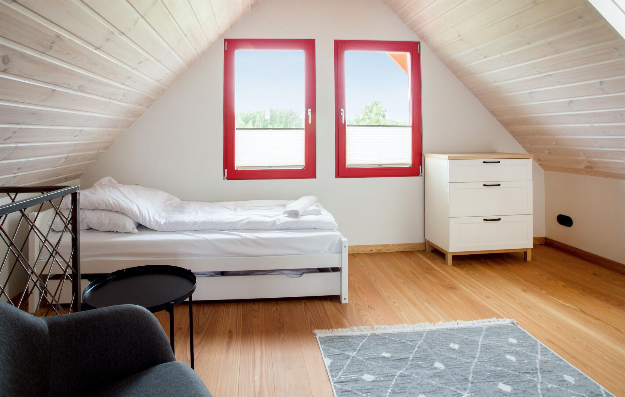 Un petit dormitori sota els teulats, ben il·luminat, amb un llit, una petita còmoda i dues finestres amb marc vermell.