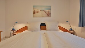 Ein Doppelbett mit weißer Bettwäsche, zwei Kissen, ein hölzernes Kopfende und zwei Nachttische mit Lampen. An der Wand hängt ein Gemälde von einer verschneiten Landschaft.
