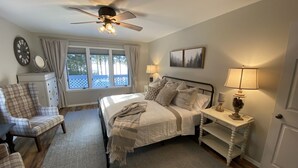 Ein Schlafzimmer mit einem Bett, zwei Nachttischen, einem Sessel, einem Deckenventilator, einem Fenster mit Vorhängen und einer Wanduhr.
